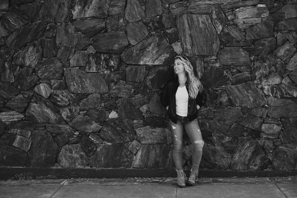 the laughing survivor leans against a rock wall wearing a leather jacket white tee and jeans smiling at someone off to the left